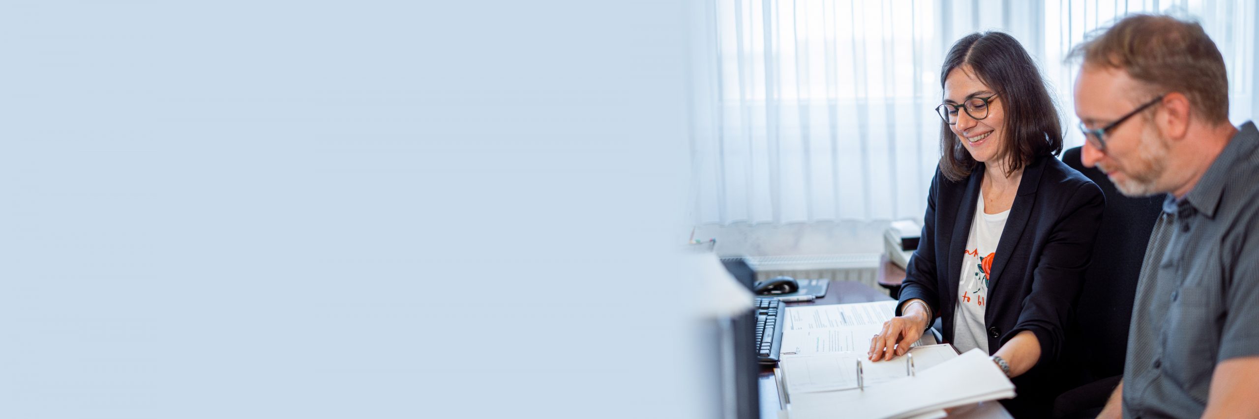 Zwei Personen sitzen an ei-nem Schreibtisch und be-trachten gemeinsam Dokumen-te. Eine Person trägt ein dunkles Sakko und eine Bril-le, die andere ein kurzärme-liges Hemd. Auf dem Schreib-tisch stehen ein Monitor, eine Tastatur sowie ver-schiedene Papiere und Büro-materialien.