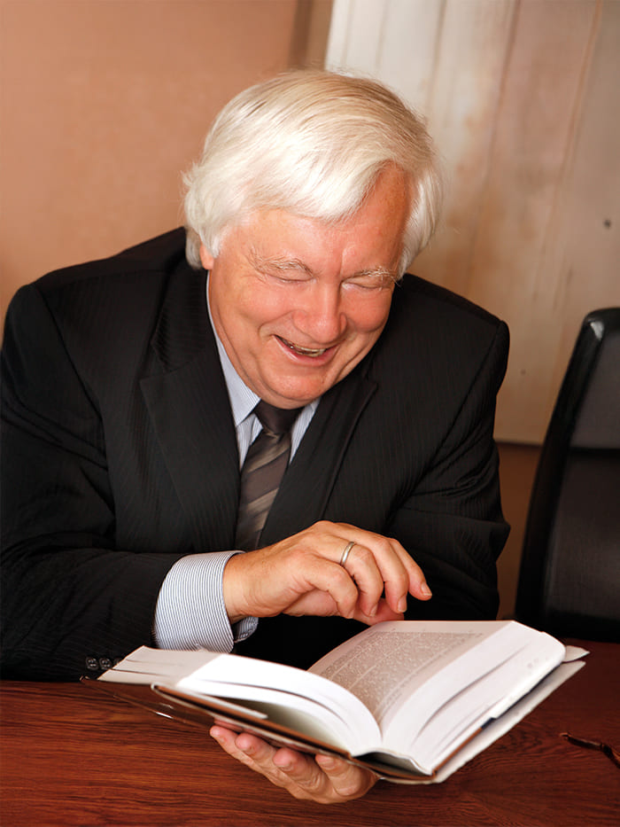 Eine Person mit weißen Haa-ren sitzt an einem Schreib-tisch und trägt einen dunk-len Anzug mit Krawatte. Vor der Person liegt ein aufge-schlagenes Buch, das sie be-trachtet.