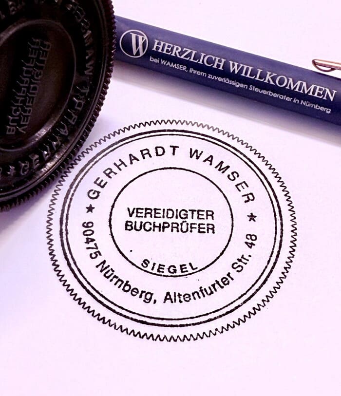 Nahaufnahme eines Stempelab-drucks auf kariertem Papier. Der Abdruck zeigt den Text: ‚WAMSER Steuerberatungsge-sellschaft mbH. Ein Teil des Stempels sowie ein Stift sind ebenfalls im Bild zu sehen.