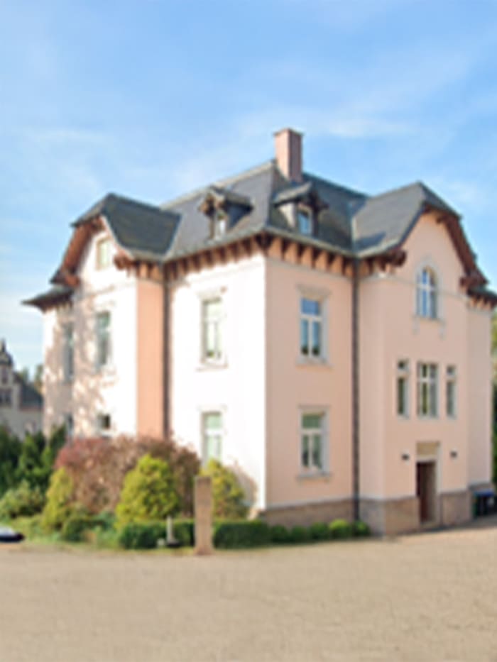 Ein großes, helles, dreistöckiges Haus mit dunkelgrauem Dach. Das Gebäude verfügt über zahlreiche Fenster auf jeder Etage sowie dekorative architektonische Elemente unter dem Dach. Umgeben ist das Haus von Büschen und Bäumen. Der Himmel im Hintergrund ist klar und blau.