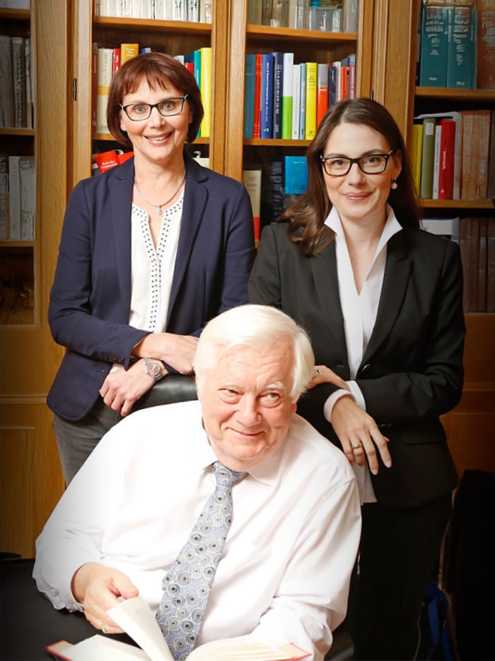 Drei Personen befinden sich in einem professionellen Um-feld, vermutlich einem Büro. Sie tragen Businesskleidung. Eine Person sitzt im Vorder-grund und hält ein aufge-schlagenes Buch, während zwei weitere Personen dahin-terstehen. Im Hintergrund ist ein Bücherregal mit zahlreichen Büchern zu se-hen.