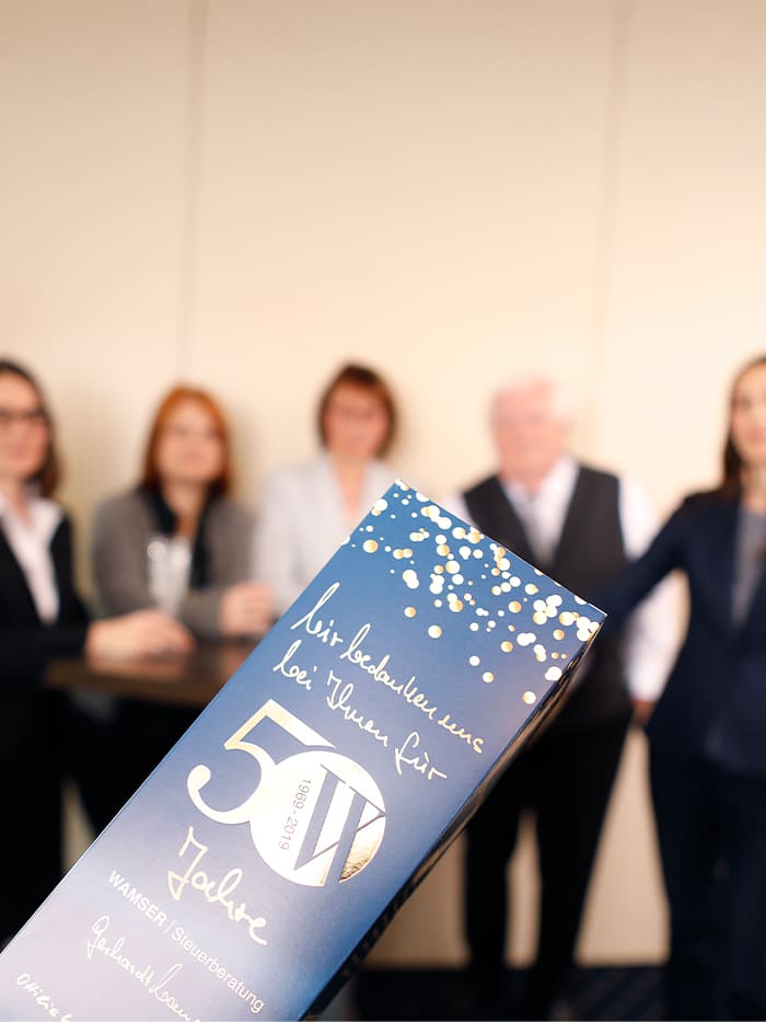 Fünf Personen stehen zusam-men. Im Vordergrund ist eine Präsent-Box in Form eine Sektflasche zu sehen. Auf der Verpackung der Sektfla-sche ist folgender Text zu erkennen: ‚Eine Einladung zum Tag der offenen Tür – 50 Jahre Jubiläum‘.