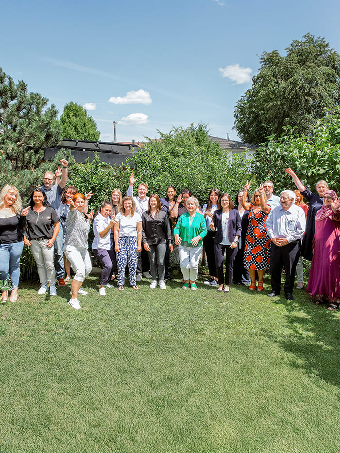 Eine Gruppe von etwa 18 Per-sonen steht im Freien vor grüner Vegetation und po-siert für ein Gruppenfoto. Einige heben die Arme oder machen Gesten, was auf einen feierlichen Moment hindeu-tet. Die Personen tragen Kleidung von lässig bis halbformell in verschiedenen Farben und Stilen.