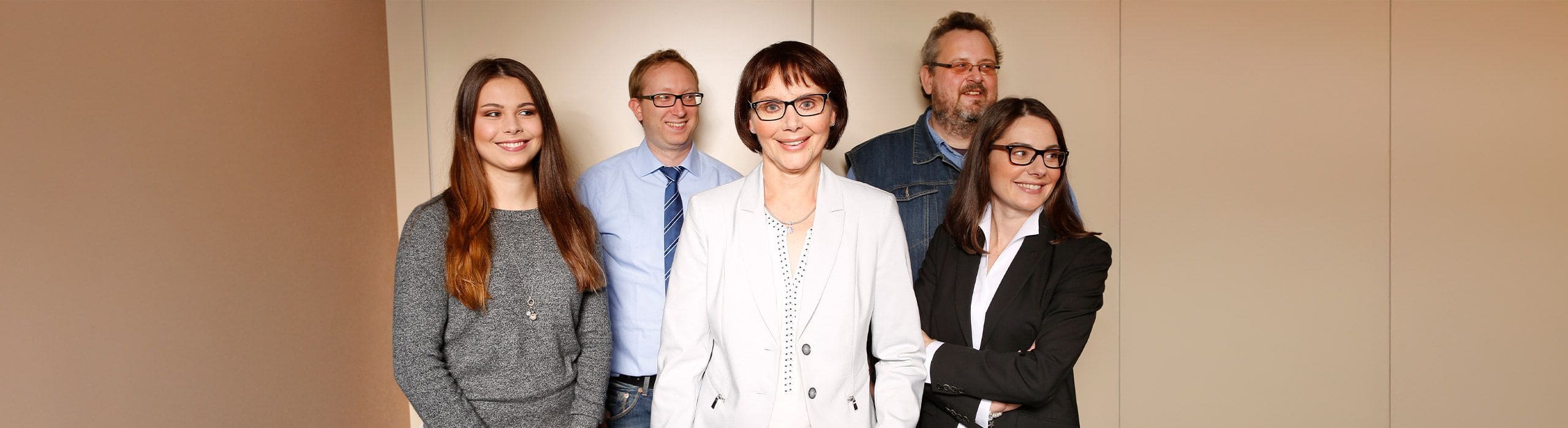 Gruppe von ungefähr 20 Per-sonen steht bei Tageslicht auf einer Wiese vor einer Hecke und Bäumen. Die Perso-nen tragen unterschiedliche Kleidung – von Freizeit- bis formeller Kleidung – und stehen dicht beieinander. Einige heben die Arme oder machen Gesten.