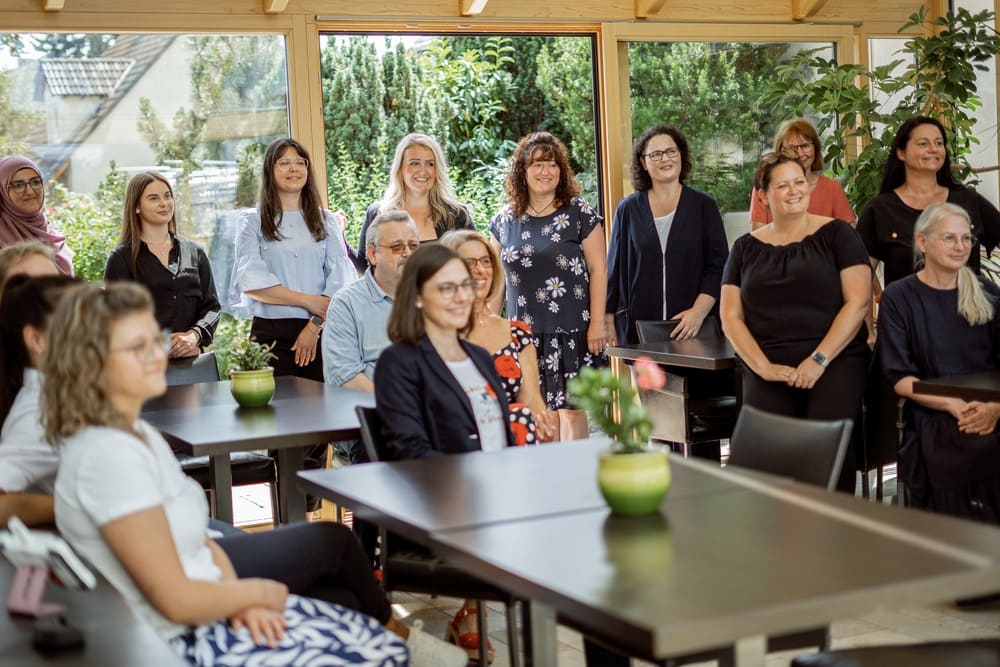 Gruppe von Personen in einem Raum mit großen Fenstern. Draußen ist Grün zu sehen. Mehrere Tische mit grünen Topfpflanzen stehen im Raum. Die Personen stehen und sit-zen um die Tische und bli-cken nach vorn.