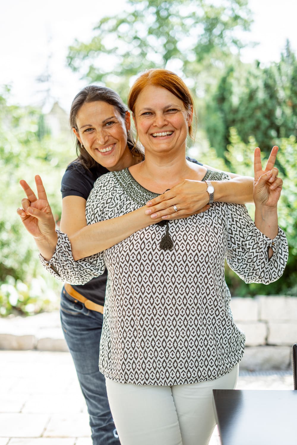 Zwei Personen stehen im Freien. Eine Person steht hinter der anderen und legt die Arme um deren Schultern. Eine Person zeigt mit den Händen das Peace-Zeichen. Im Hintergrund sind Grünflächen und eine Steinmauer zu se-hen.