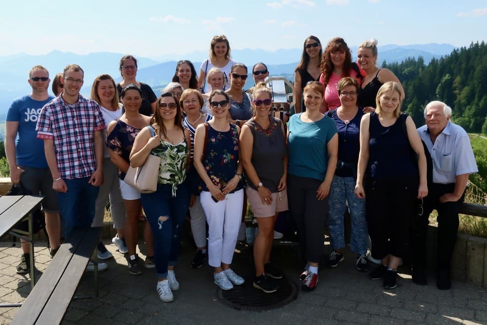 Gruppe von etwa 20 Personen posiert im Freien für ein Foto. Im Hintergrund sind Berge und Bäume zu sehen. Einige Personen stehen, an-dere sitzen auf Bänken.