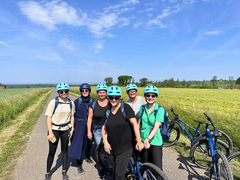 Sechs Personen stehen auf einem gepflasterten Weg in ländlicher Umgebung, umgeben von Feldern. Sie tragen alle Fahrradhelme und neben ihnen stehen Fahrräder, was auf eine Radtour hinweist. Der Himmel ist leicht bewölkt und sonnig.