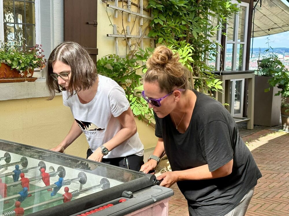 Zwei Personen spielen im Freien Tischfußball. Der Ort wirkt wie eine Terrasse, im Hintergrund sind Pflanzen und Grün zu sehen.