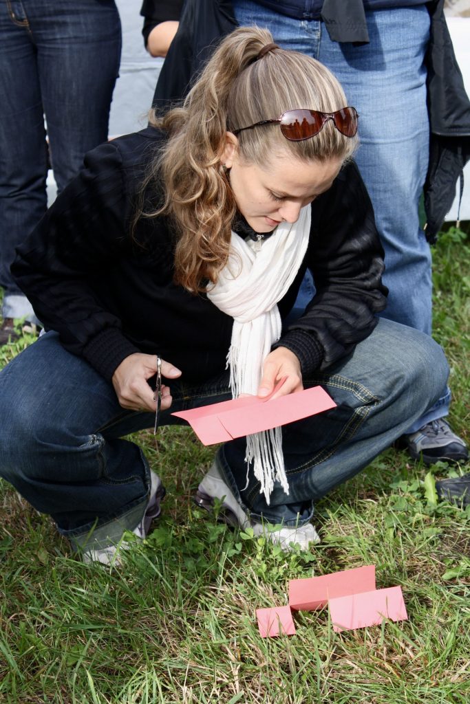 	Eine Person mit langen Haaren und Sonnenbrille kniet auf einer Wiese und schneidet ein rosa Papierstück mit einer Schere. Vor ihr liegen bereits mehrere ausgeschnittene Papierstücke. Im Hintergrund sind weitere Menschen zu sehen.