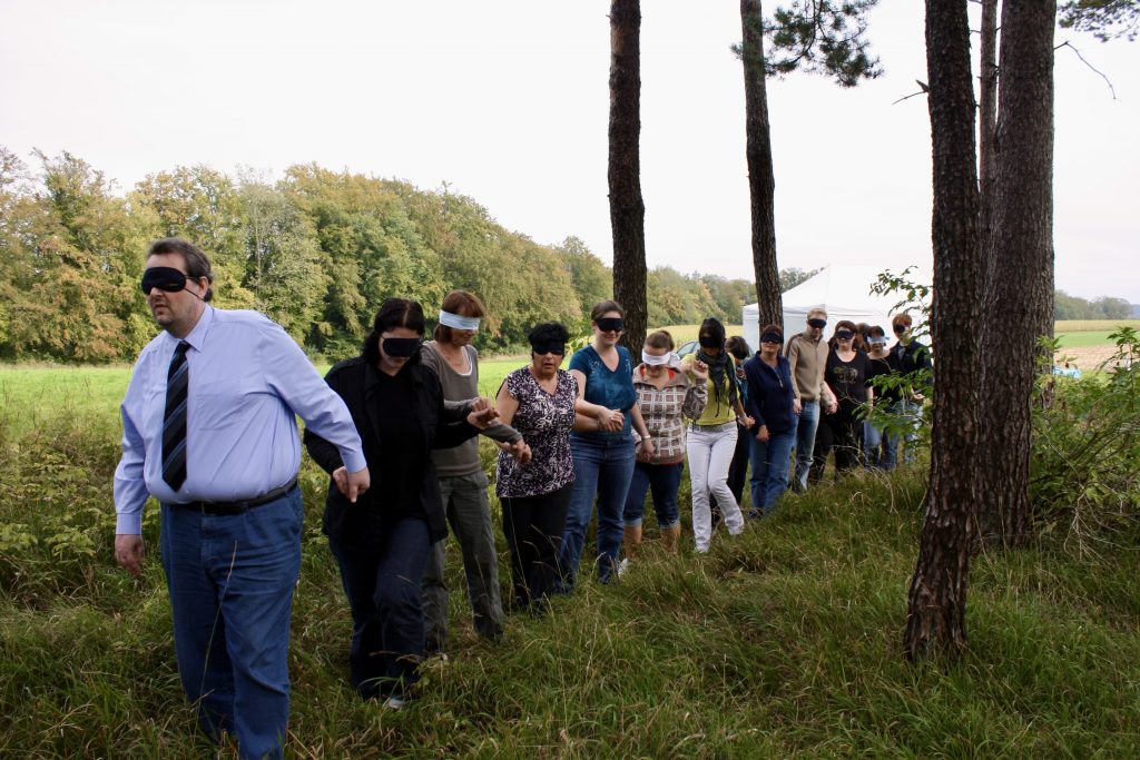 Eine Gruppe von Menschen geht in einer Reihe durch eine grasbewachsene Fläche mit Bäumen. Die meisten tra-gen Augenbinden und halten sich zur Orientierung an der Person vor ihnen fest. Die Szene spielt im Freien, in einem Waldgebiet.