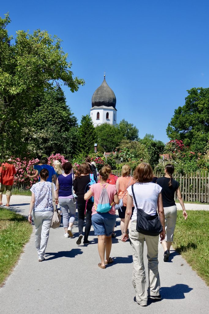 Eine Gruppe von Menschen geht auf einem gepflasterten Weg durch eine grüne Landschaft mit Bäumen und Gras. Rechts ist ein Holzzaun zu sehen, im Hintergrund steht ein Gebäude mit einem kuppelartigen Dach. Der Himmel ist klar und blau.