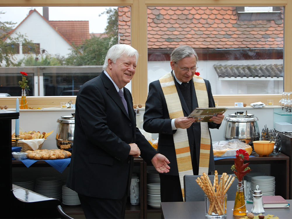 Wamser Zitat Zwei Personen stehen vor einem Buffet-Tisch mit verschiedenen Speisen und Utensilien. Eine Person hält ein Buch. Beide sind formell ge-kleidet – eine im Anzug, die andere in religiöser Kleidung mit Stola. Im Hintergrund sind Fenster mit Blick auf Dächer und etwas Grün zu sehen.