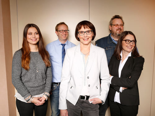 Fünf Personen stehen zusam-men und posieren für ein Gruppenfoto vor einem schlichten Hintergrund. Die Kleidung reicht von Busi-ness- bis Freizeitkleidung, darunter Blazer, Pullover und Hemden.
