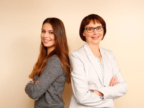 Zwei Personen stehen Rücken an Rücken. Die linke Person hat lange braune Haare und trägt ein graues Oberteil. Die rechte Person hat kurze braune Haare, trägt eine Brille sowie einen hellen Blazer über einem weißen Hemd mit kleinen schwarzen Punkten.