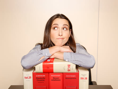 Eine Person sitzt hinter ei-nem Stapel großer Bücher. Die Person trägt ein ge-streiftes Oberteil. Die Bü-cher sind überwiegend rot und weiß und tragen Titel wie „Deutsche Gesetze“, „Steuergesetze“ und „HGB“.